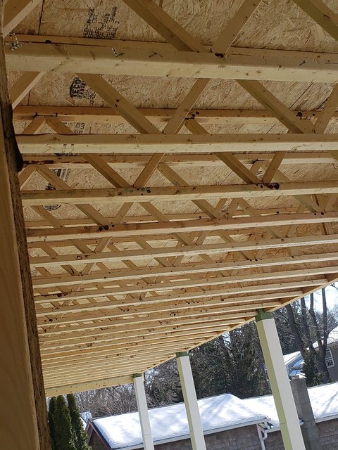 Underside of a newly framed deck showing open-web wooden joists and white support posts in a snowy backyard