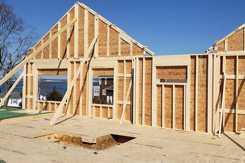 Framed gable walls and window openings on a second floor, with a bay and distant shoreline visible through the openings under a clear blue sky