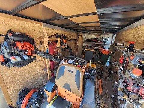 Inside the equipment trailer: an orange Scag mower in the middle, backpack blowers on one wall, string trimmers on racks on the other, and a Fro's Landscape and Construction sign at the back